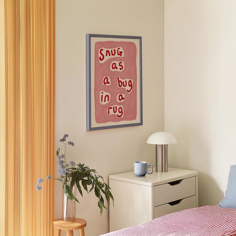 May include: A framed art print with the phrase "Snug as a bug in a rug" in red letters on a pink background. The artwork hangs on a cream-colored wall above a nightstand with a lamp and a blue mug.