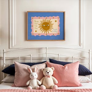 Puede incluir: Un cuadro enmarcado con una ilustraci&oacute;n de un sol sonriente y el texto "Here Comes The Sun Little Barley" cuelga sobre una cama con almohadas rosas y peluches. La obra de arte tiene un borde azul y un marco interior rosa.