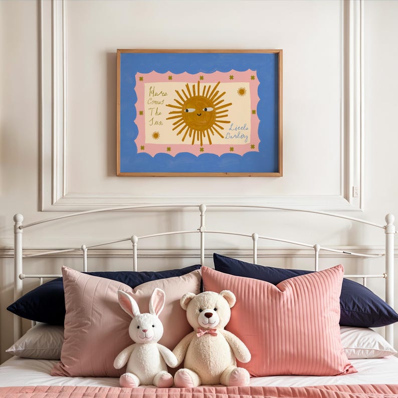 Puede incluir: Un cuadro enmarcado con una ilustraci&oacute;n de un sol sonriente y el texto "Here Comes The Sun Little Barley" cuelga sobre una cama con almohadas rosas y peluches. La obra de arte tiene un borde azul y un marco interior rosa.