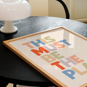 Puede incluir: Una impresi&oacute;n enmarcada con el texto "This Must Be The Place" en letras de colores sobre un fondo crema. El marco es de madera clara y la impresi&oacute;n est&aacute; sobre una mesa negra.