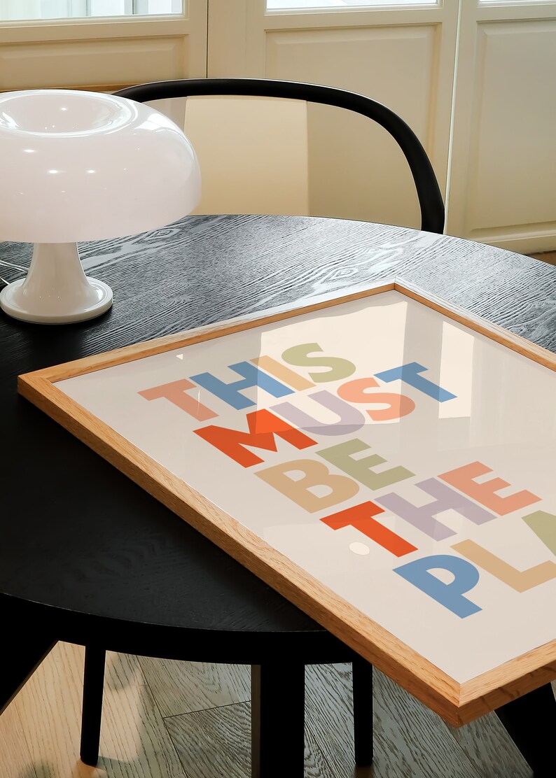 Puede incluir: Una impresi&oacute;n enmarcada con el texto "This Must Be The Place" en letras de colores sobre un fondo crema. El marco es de madera clara y la impresi&oacute;n est&aacute; sobre una mesa negra.