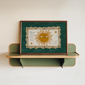 Puede incluir: Una impresi&oacute;n art&iacute;stica enmarcada con una ilustraci&oacute;n de un sol sonriente y el texto "Here Comes The Sun Little Darling". La obra de arte tiene un borde verde y se exhibe en una estanter&iacute;a verde y de madera.