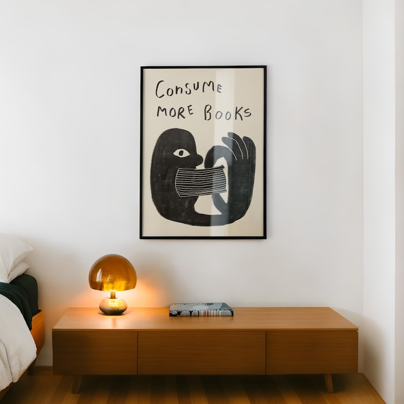 May include: A black and white illustration of a person consuming books with the text "Consume More Books" in a black frame hanging on a white wall above a wooden dresser with a lamp and a book on top.