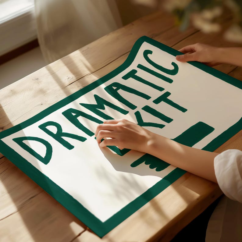 Puede incluir: Un cartel blanco con un borde verde y las palabras "Dramatic Kit" escritas en negro. El cartel est&aacute; sobre una superficie de madera.
