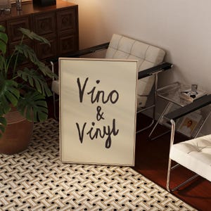 Puede incluir: Un cuadro enmarcado con las palabras "Vino & Vinyl" en escritura negra. El cuadro est&aacute; en un marco de color claro y est&aacute; apoyado contra una pared. La imagen est&aacute; en una habitaci&oacute;n con una alfombra estampada y muebles modernos.