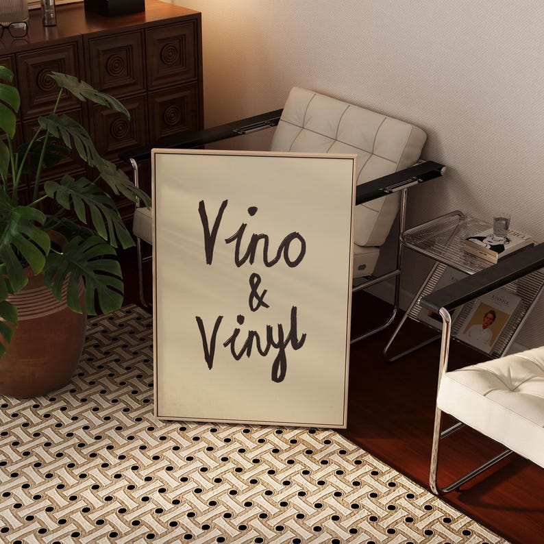 Puede incluir: Un cuadro enmarcado con las palabras "Vino & Vinyl" en escritura negra. El cuadro est&aacute; en un marco de color claro y est&aacute; apoyado contra una pared. La imagen est&aacute; en una habitaci&oacute;n con una alfombra estampada y muebles modernos.