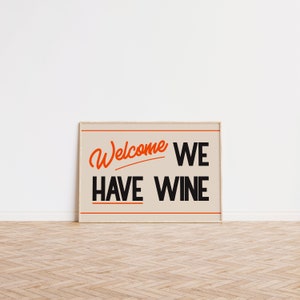 May include: A beige framed sign with the text "Welcome We Have Wine" in black and orange lettering.