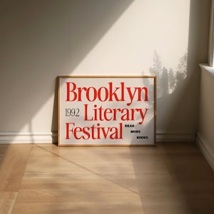 May include: A framed poster with the words "Brooklyn Literary Festival 1992" in red, with the text "Read More Books" at the bottom. The poster is on a light-coloured wooden floor, leaning against a white wall.