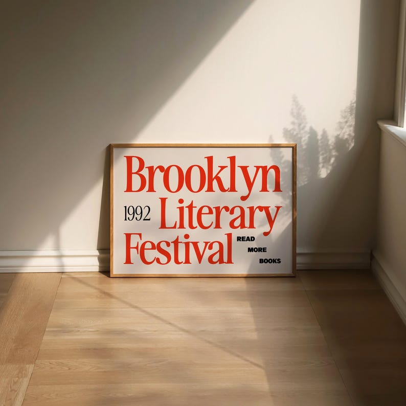 May include: A framed poster with the words "Brooklyn Literary Festival 1992" in red, with the text "Read More Books" at the bottom. The poster is on a light-coloured wooden floor, leaning against a white wall.