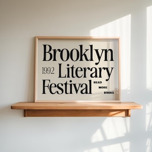 May include: A framed poster with the text "Brooklyn Literary Festival 1992" in black font. The poster is displayed on a wooden shelf against a white wall. The phrase "Read More Books" is also included.