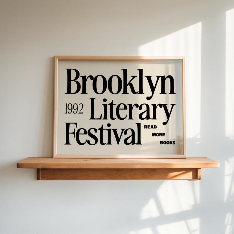 May include: A framed poster with the text "Brooklyn Literary Festival 1992" in black font. The poster is displayed on a wooden shelf against a white wall. The phrase "Read More Books" is also included.