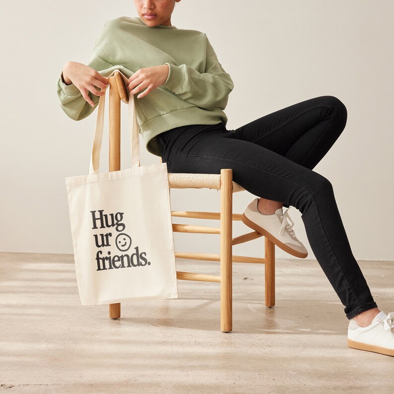 May include: A white tote bag with black text that reads "Hug ur friends." and a smiley face. The bag is being held by a person wearing a light green sweater and black jeans.