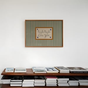 K&ouml;nnte beinhalten: Ein gerahmtes Kunstwerk mit gr&uuml;n-wei&szlig; gestreiftem Hintergrund und einem zentralen Rechteck mit dem Text "Night Night Love You" und blauen Sternakzenten. Das Kunstwerk wird &uuml;ber einem Holzregal mit mehreren B&uuml;chern ausgestellt.