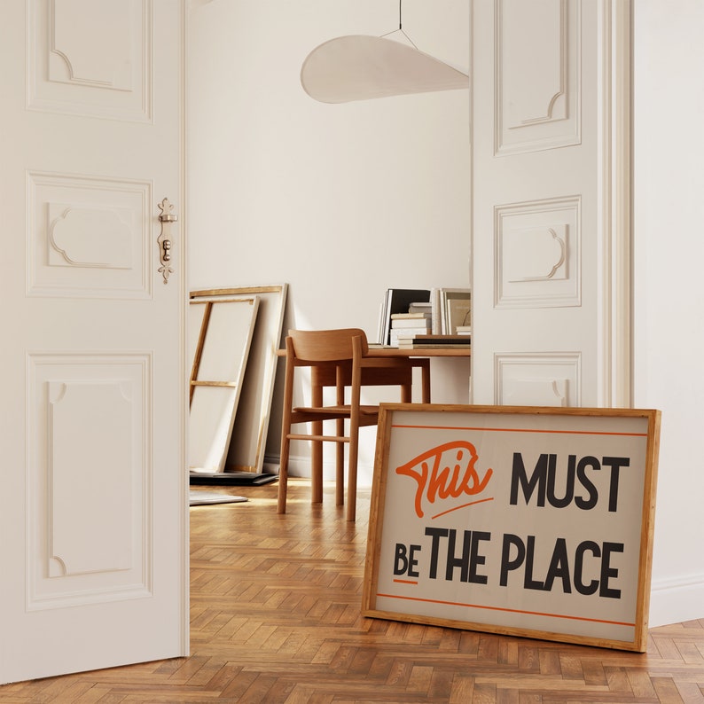 May include: A framed print with the text "This Must Be The Place" in orange and black lettering on a beige background. The print is hanging on a wall in a room with wooden floors.