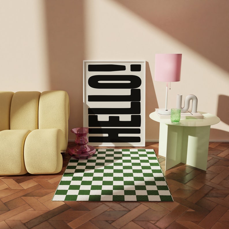 May include: A black and white poster with the word "HELLO!" in bold letters hangs on a wall above a green and white checkered rug. A yellow couch sits in front of the rug. A pink lamp with a white shade sits on a light green side table.