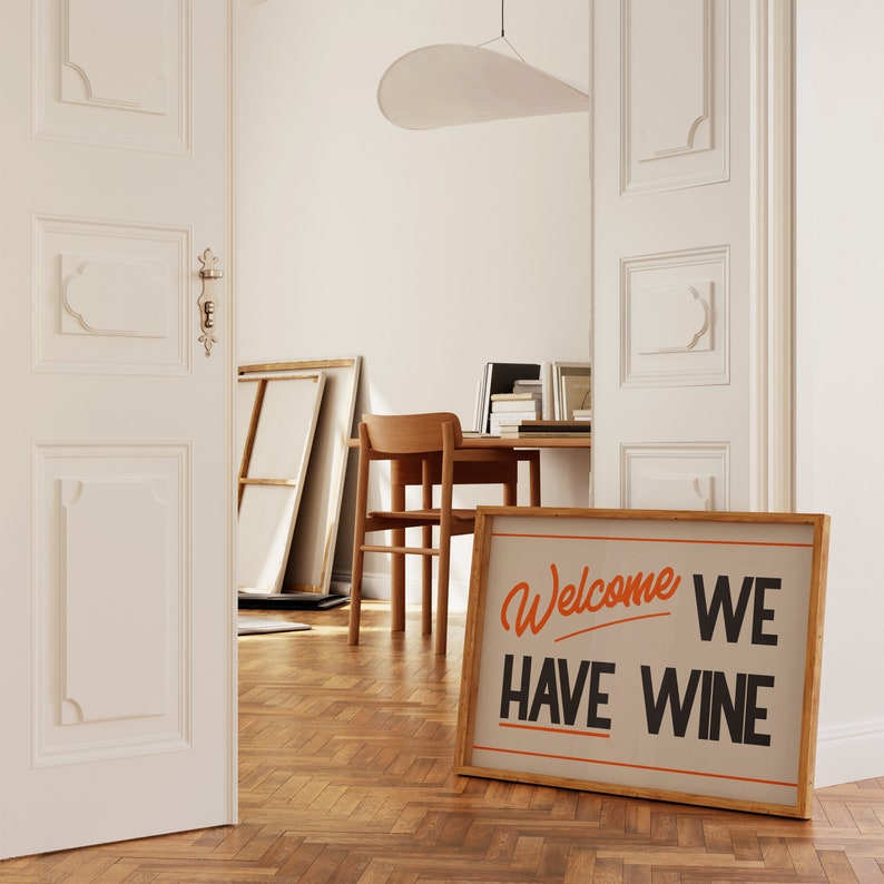May include: A framed sign with the text "Welcome We Have Wine" in orange and black lettering on a beige background. The sign is leaning against a wooden floor.