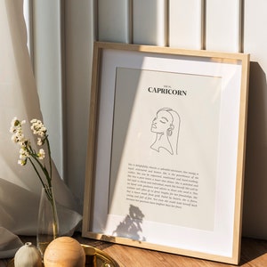 May include: A framed print with a minimalist line drawing of a woman's face and the text "SHE IS CAPRICORN" on a white background. The frame is light brown wood and the print is sitting on a wooden surface.