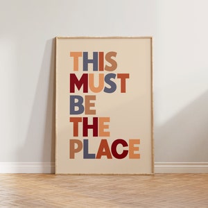 Puede incluir: Una impresi&oacute;n enmarcada con el texto "THIS MUST BE THE PLACE" en un dise&ntilde;o de fuente colorido y en capas. Las letras est&aacute;n en tonos de rojo, naranja, marr&oacute;n y azul.