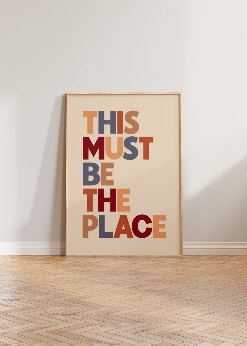 Puede incluir: Una impresi&oacute;n enmarcada con el texto "THIS MUST BE THE PLACE" en un dise&ntilde;o de fuente colorido y en capas. Las letras est&aacute;n en tonos de rojo, naranja, marr&oacute;n y azul.