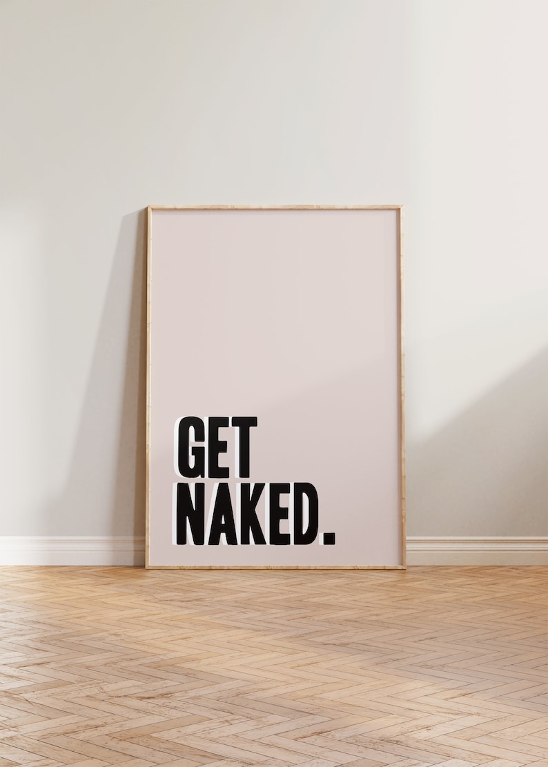 May include: A rectangular wall art print in a light wooden frame featuring a pale pink background with the text GET NAKED. printed in bold black capital letters. The artwork is displayed in a vertical orientation with a clean, simple design.