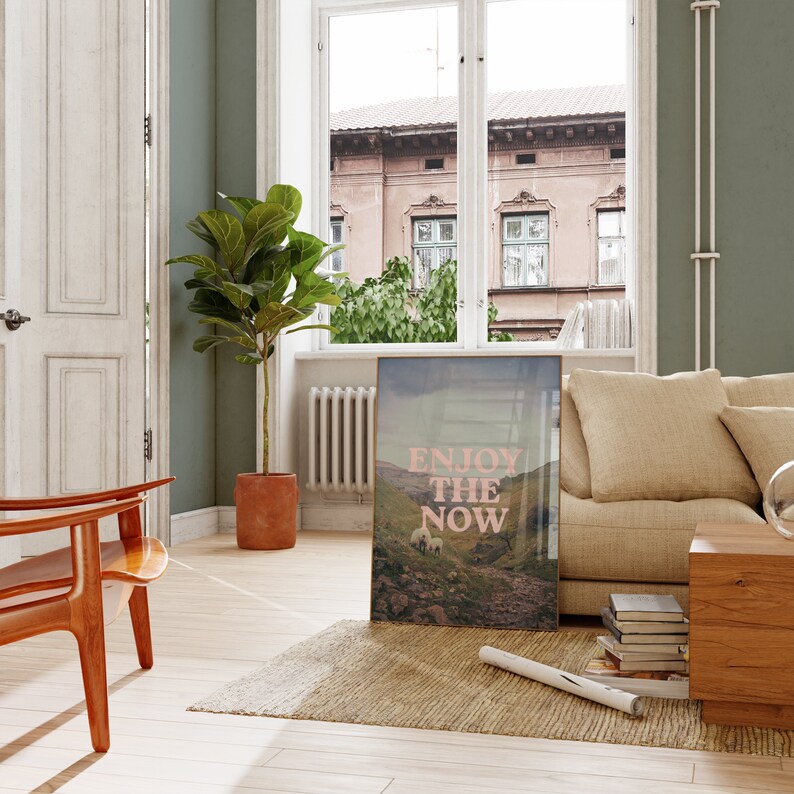 以下が含まれることがあります： 緑の丘陵の風景が描かれた額装されたプリントで、ピンクのテキスト「Enjoy the Now」が重ねられています。プリントは、ソファ、椅子、植物のあるリビングルームの壁に飾られています。