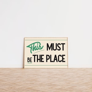 May include: A framed print with a white background and black text that reads "This Must Be The Place" in a retro font. The word "This" is written in green.