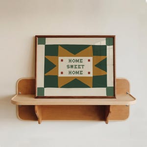 Puede incluir: Una impresi&oacute;n rectangular enmarcada con un patr&oacute;n de estrellas en verde oscuro y amarillo mostaza sobre un fondo crema. El centro contiene el texto "HOME SWEET HOME" en letras may&uacute;sculas verde oscuro, flanqueado por cuatro estrellas rojas.