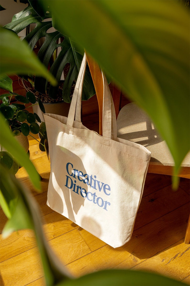 May include: A beige canvas tote bag with blue text that reads "Creative Director". The bag is sitting on a wooden floor with a green plant in the background.