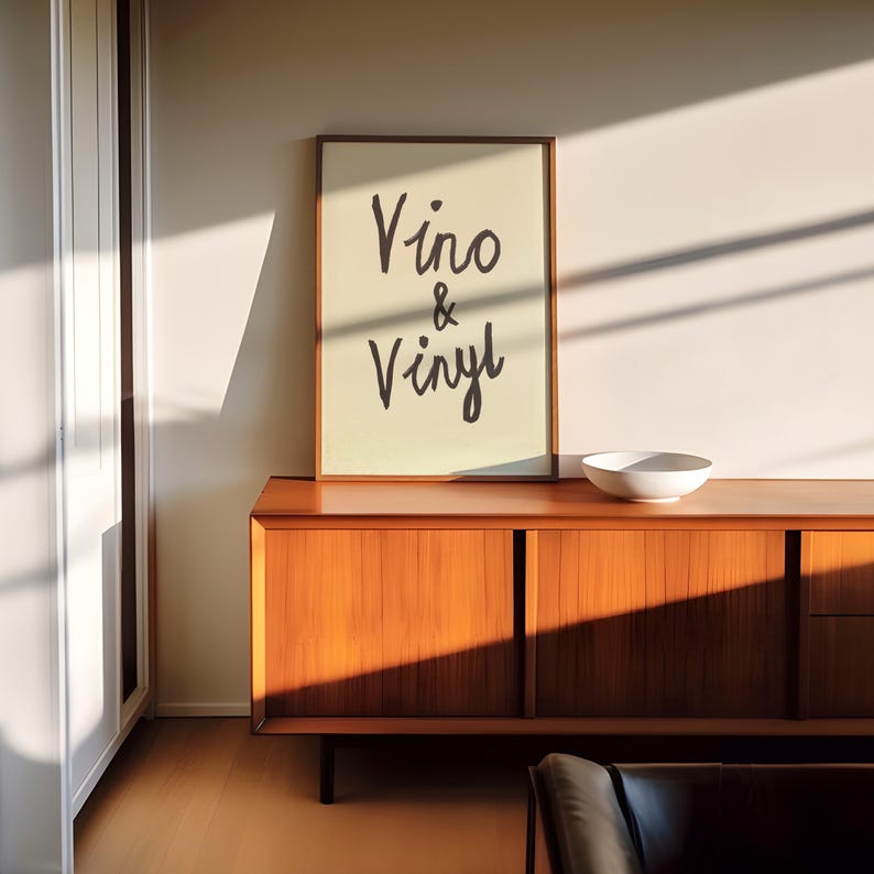 Puede incluir: Un cuadro enmarcado con las palabras "Vino & Vinyl" en escritura negra. El cuadro se exhibe sobre un mueble de madera con un cuenco blanco. El mueble est&aacute; en una habitaci&oacute;n con luz natural.