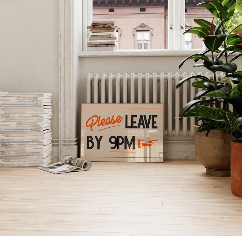 May include: A rectangular sign with the words "Please Leave By 9PM" in orange and black. The sign is propped against a wall near a window, with plants and a stack of magazines nearby.