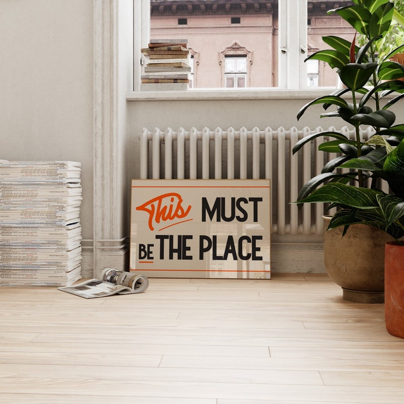 May include: A beige poster with black text that reads "This MUST BE THE PLACE" in a retro font. The word "This" is in orange. The poster is hanging on a white wall in a room with a window and a radiator.