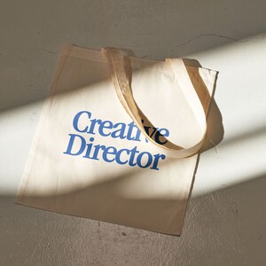 May include: A beige canvas tote bag with blue text that reads "Creative Director".