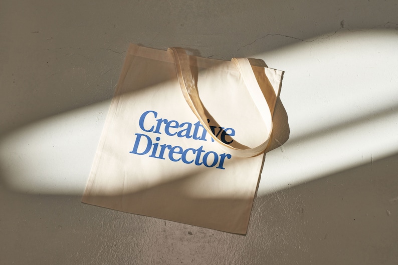 May include: A beige canvas tote bag with blue text that reads "Creative Director".