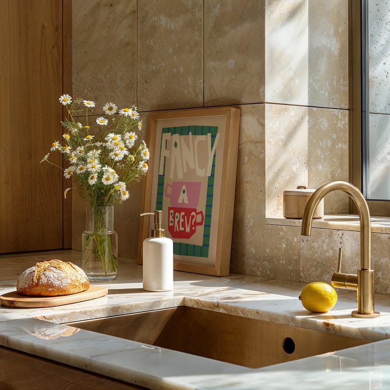 Op de afbeelding: Een gootsteen in een keuken met een gouden kraan, een witte zeepdispenser, een brood op een houten snijplank, een vaas met witte bloemen en een ingelijst print met de tekst "Fancy a Brew?"