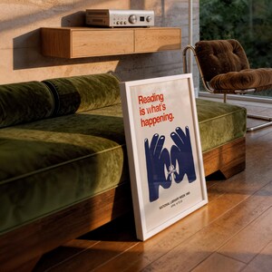 May include: A framed poster with the text "Reading is what's happening" in red, with blue hand illustrations. The poster is leaning against a green velvet sofa. The room has a wooden floor and a brown chair.