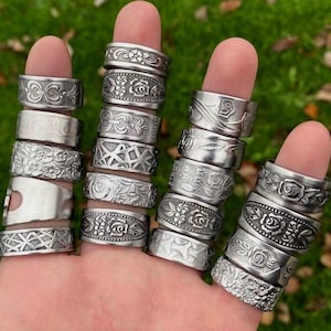 May include: An assortment of silver rings, each with a unique embossed design, including flowers and geometric shapes. The rings are displayed on fingers, highlighting the different patterns. The rings are made of metal and have a vintage or handmade appearance.