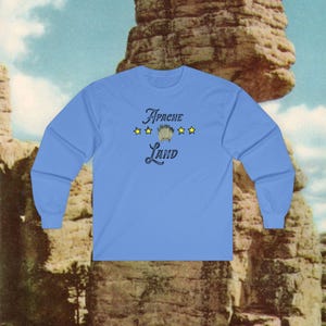 May include: A light blue long-sleeve shirt with the text "Apache Land" in a stylized font. The design includes three gold stars and a Native American headdress graphic. The shirt is made of a soft material.