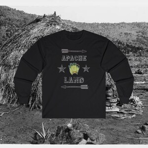 Può includere: Maglietta nera a maniche lunghe con un design grafico bianco che presenta un copricapo nativo americano e il testo "Apache Land" con due stelle su entrambi i lati del copricapo.