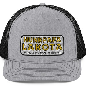 Puede incluir: Gorra de camionero gris con la parte trasera de malla negra. La parte delantera presenta un parche bordado con las palabras "HUNKPAPA LAKOTA" en letras doradas y "NATIVE LANDS CLOTHING COMPANY" debajo.