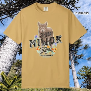 Puede incluir: Camiseta de color mostaza con una ilustración detallada de un coyote y el texto "SIERRA MIWOK TRIBE CALIFORNIA". El diseño incluye detalles florales. La camiseta está etiquetada como "10 COLORES".