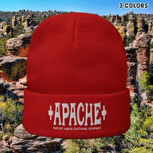 May include: A red beanie with the word "APACHE" in white, along with "NATIVE LANDS CLOTHING COMPANY" below. The text "3 COLORS" is in the top right corner. The background features a natural landscape with rock formations and a blue sky.