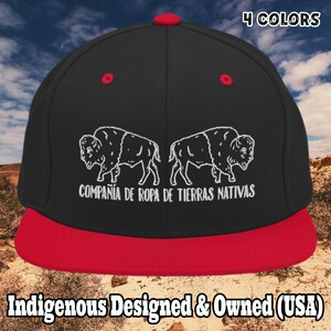 May include: Black and red flat-brimmed cap with a white bison graphic and text. The cap features the text "COMPAÑÍA DE ROPA DE TIERRAS NATIVAS" and "Indigenous Designed & Owned (USA)". The cap is available in 4 colors.