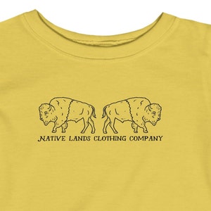 Può includere: Una maglietta gialla con il contorno nero di due bisonti che si fronteggiano. Il testo "NATIVE LANDS CLOTHING COMPANY" è stampato sotto i bisonti.
