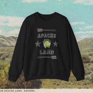 Può includere: Felpa nera con un design grafico bianco che dice "Apache Land" con un copricapo nativo americano e due frecce.