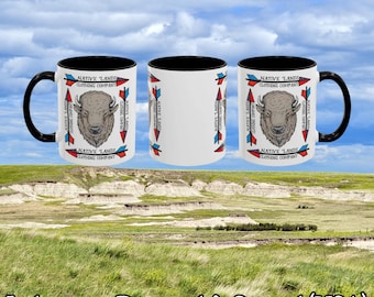 May include: White ceramic mug with a black handle and interior. The mug features a bison illustration, red and blue arrows, and the text "NATIVE LANDS CLOTHING COMPANY". The mug is set against a landscape with a cloudy sky.