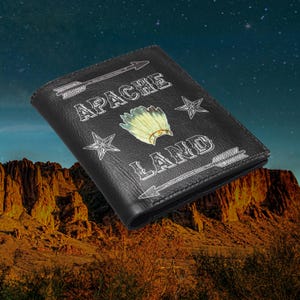 May include: Black leather wallet with the words "APACHE LAND" in white lettering, along with arrows and star designs. A Native American headdress is in the center. The wallet is rectangular and appears to be closed.