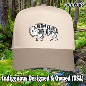 May include: A beige and brown baseball cap featuring a black graphic of a bison with the text "NATIVE LANDS CLOTHING COMPANY." The cap has a curved brim and a structured crown. The text "3 COLORS" is visible on the top right. The text "Indigenous Designed & Owned (USA)" is visible at the bottom.