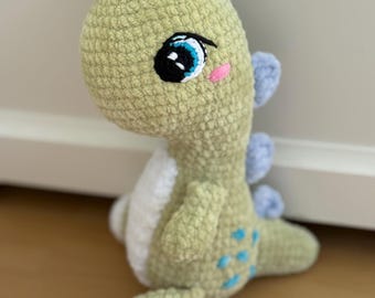 May include: A handmade, plush dinosaur toy in shades of green, white, and blue. The crochet dinosaur has large, expressive eyes, a pink cheek, and blue accents on its back and head. The toy is sitting on a wooden surface.