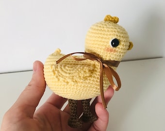 May include: A handmade crochet chick in light yellow with brown legs and a brown ribbon bow. The chick has black eyes and a small yellow crest. The cheeks are lightly blushed. The chick is a cute, handcrafted item.