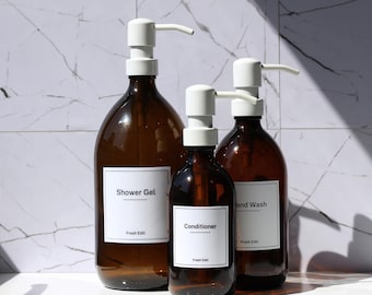 May include: Three amber glass bottles with white pump dispensers. The bottles are labeled "Shower Gel", "Conditioner", and "Hand Wash" with the brand "Fresh Edit". The bottles are arranged in front of a white marble tile background.